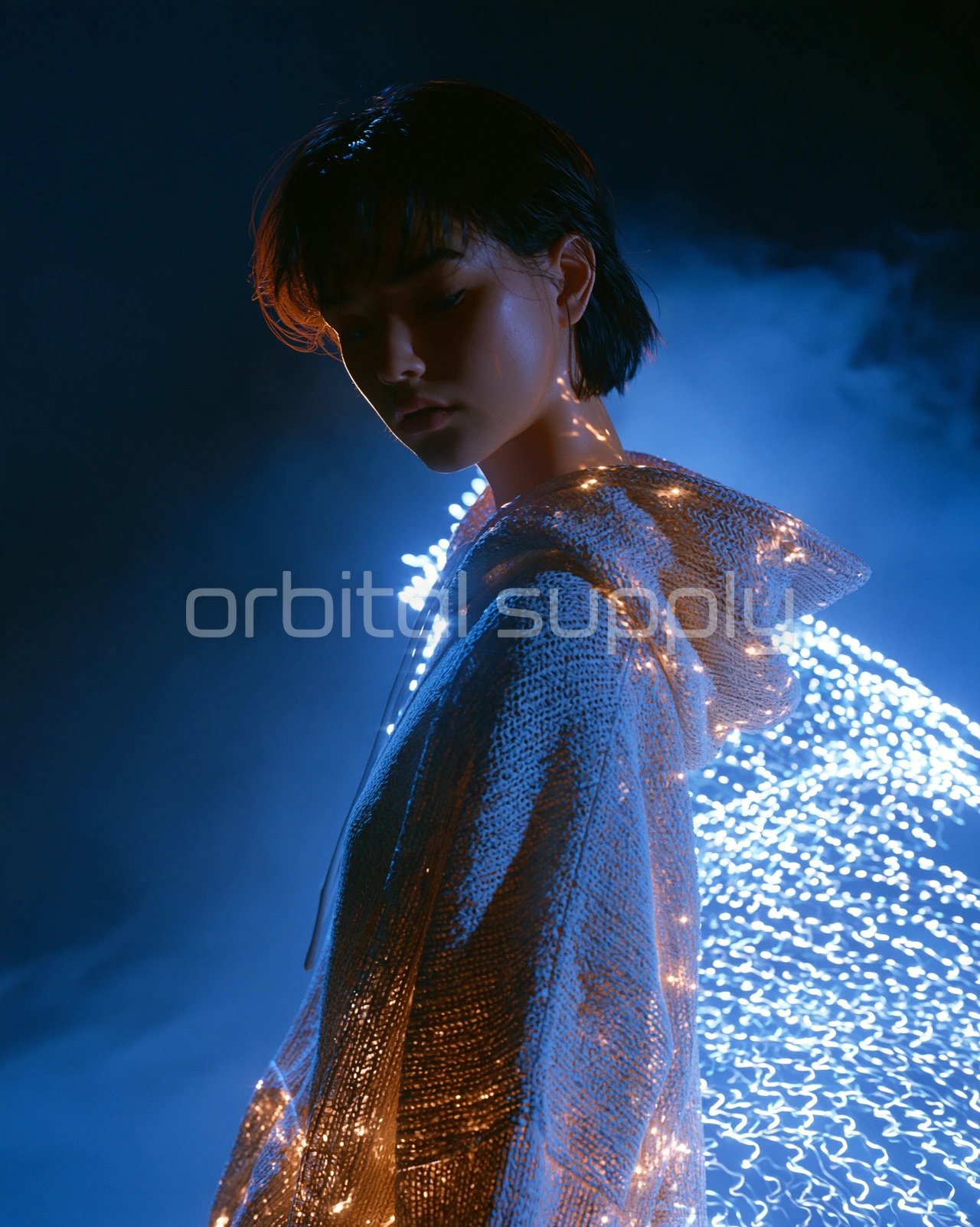 A young person with short dark hair looks down, the white hoodie lit by cold blue light and small gold reflections.